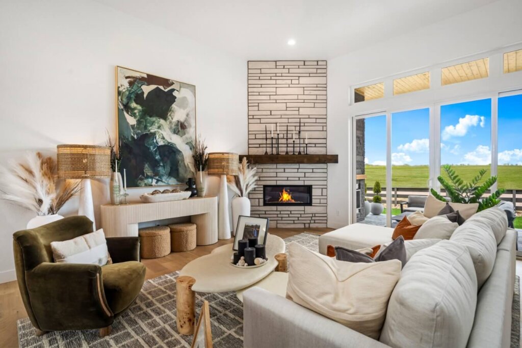 A modern living room with a lit fireplace, large abstract painting, and neutral-toned furniture. Large windows reveal a view of a grassy field and blue sky with clouds, echoing the serene landscapes seen in model homes Loveland is known for.