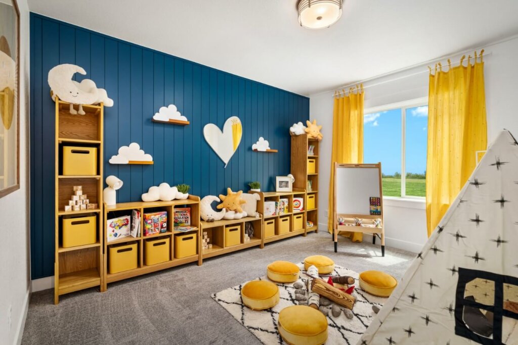 The children's playroom features a blue accent wall, yellow storage bins, cloud decorations, a small teepee, and an easel. Sunlight pours through a window with yellow curtains, creating a bright space reminiscent of the vibrant designs found in model homes Loveland showcases.