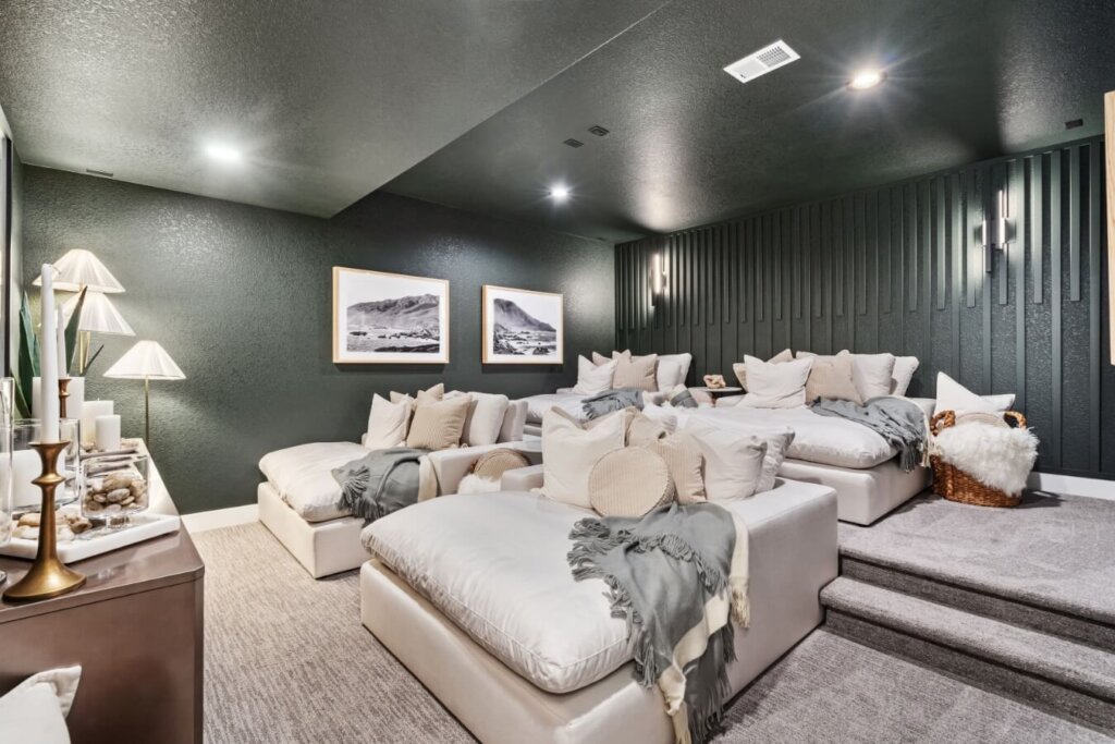 A cozy home theater in the style of model homes Loveland offers, with tiered seating featuring plush couches and pillows, dark green walls, and framed artwork. Perfect for unwinding after a delightful meal at Centerra restaurants.