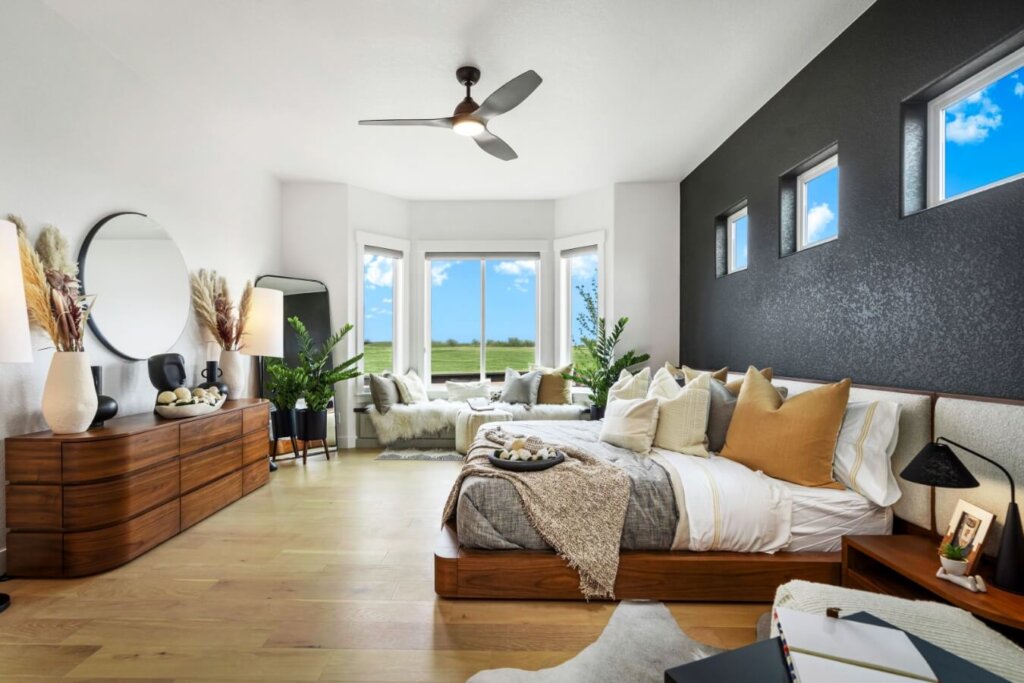 Spacious bedroom with a modern wooden bed, cozy textiles, and a large round mirror. Potted plants add warmth, while the bay window offers stunning landscape views, echoing the elegant design found in model homes Loveland is known for.