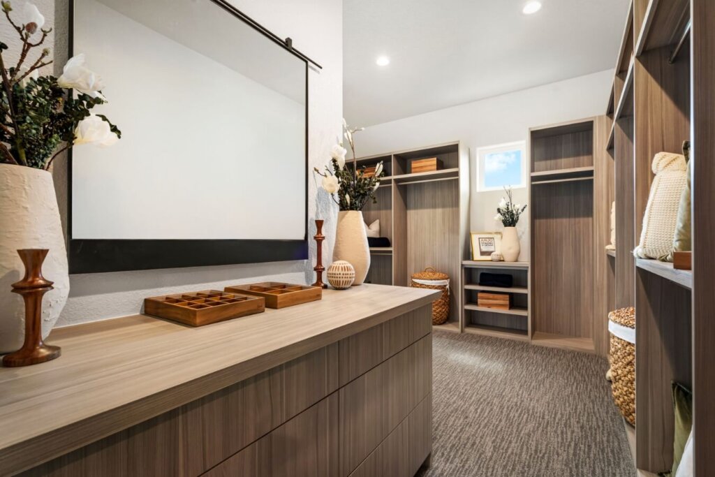 The walk-in closet, resembling the elegant designs of model homes in Loveland, features wooden shelves and drawers, a large mirror, decorative vases and baskets. A carpeted floor and overhead lighting create a cozy ambiance that echoes the charm of Kinston Centerra living.