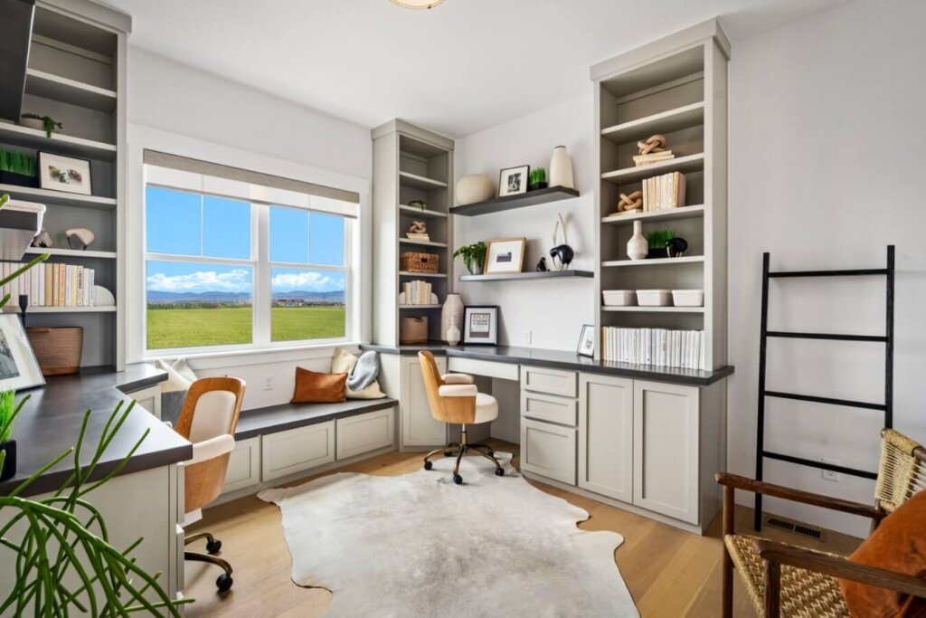 A bright home office at Kinston Centerra offers a scenic view of fields and mountains. It features built-in shelves, a desk, a chair, decorative items, and a cowhide rug on the wooden floor. Ideal for those inspired by model homes in Loveland.