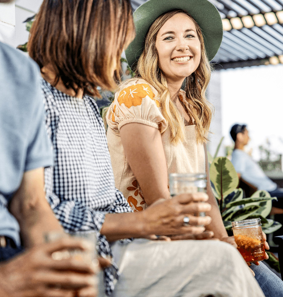 Three people sitting outside with drinks at Kinston Centerra. The woman in the center is smiling and wearing a green hat, enjoying the lively atmosphere surrounded by Centerra restaurants.