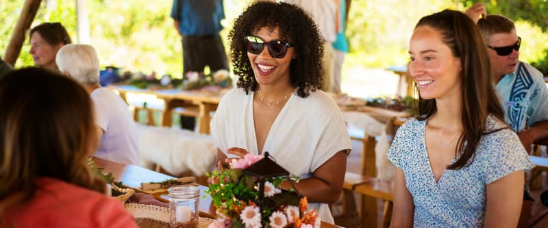 Three people are seated at an outdoor table in Centerra, Loveland Colorado, engaged in conversation. The table has floral decorations, and other people are seated at tables in the background.