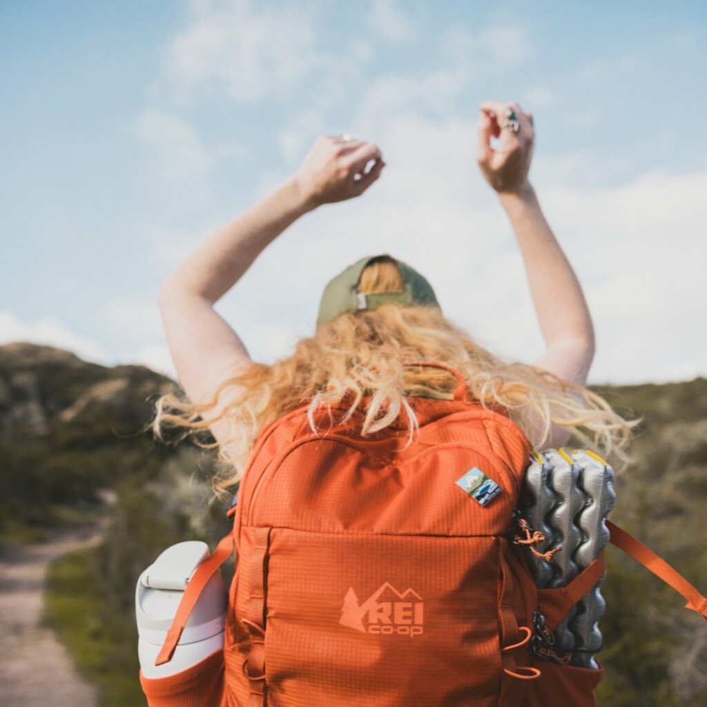 A person with long hair wearing a cap and an orange backpack, possibly from REI, raises their arms in triumph while hiking along a scenic trail near Centerra in Loveland, Colorado.