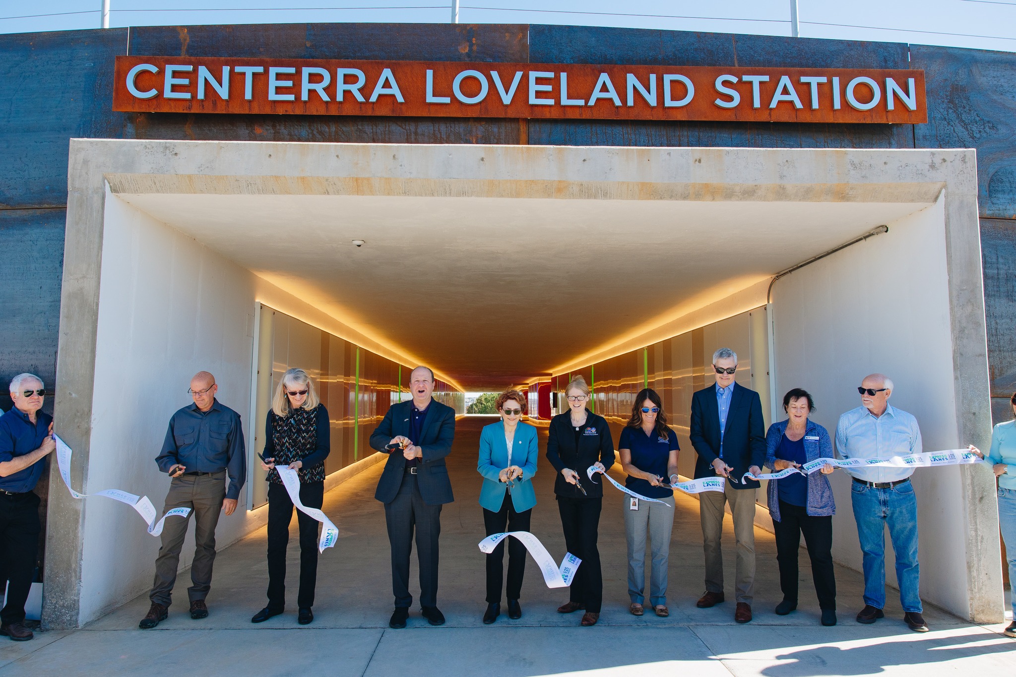 Jared Polis alongside a group of people, ceremoniously cutting a ribbon at the entrance to the Centerra Loveland Station tunnel.